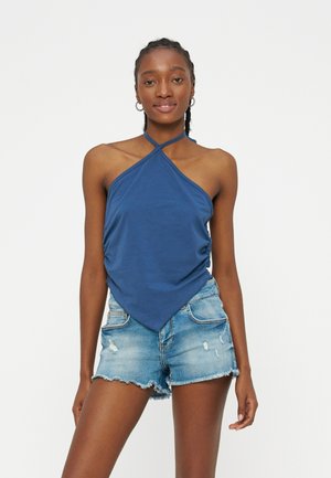 Γυναίκα που φοράει μπλε τοπ με τιράντα στο λαιμό και τριγωνικό μπροστινό μέρος, καθώς και σκισμένα τζιν σορτς, στέκεται μπροστά σε ένα απλό λευκό φόντο.