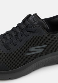 Zapatilla deportiva negra con parte superior de malla transpirable, diseño con cordones, suela de goma y acentos texturizados para agarre y soporte.
