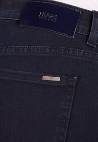 Donkerblauwe spijkerbroek met achterzak, voorzien van een klein metalen label en een zwarte leren patch, beide met het reliëf van de merknaam "HUGO".