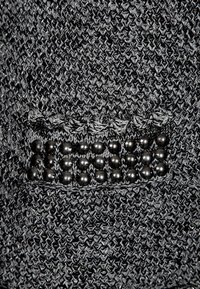 Černá pletená látka s texturovaným povrchem, s ozdobnou nášivkou zdobenou kulatými černými korálky v horizontální linii.