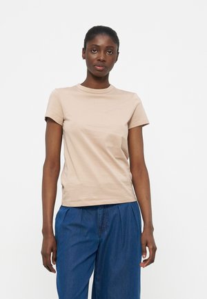 Γυναίκα που στέκεται με το πρόσωπο προς τα εμπρός, φορώντας μπεζ κοντομάνικο μπλουζάκι και μπλε φαρδιά παντελόνια, μπροστά σε ένα απλό λευκό φόντο.