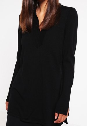 Frau trägt einen schwarzen Pullover mit langen Ärmeln, V-Ausschnitt und Seitenschlitzen, steht vor einem schlichten weißen Hintergrund.