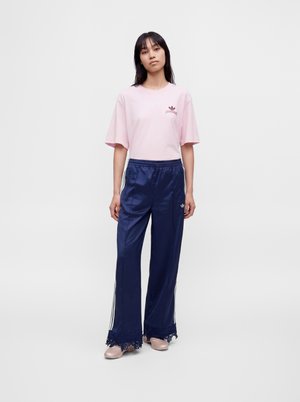 Femeie purtând un tricou roz deschis și pantaloni largi bleumarin cu dungi albe și margine de dantelă, stând în fața unui fundal simplu.