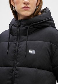 Mustapuffatakki hupulla, jossa on vetoketjusuljenta, etutaskut ja pieni logo-ompelus. Kankaan pinnassa on hieno kiilto ja rakenne.