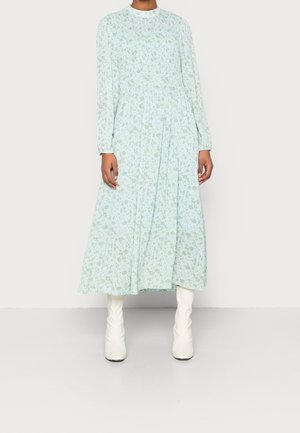 Hellblaues Blumenkleid mit hohem Ausschnitt, langen Ärmeln und plissiertem Rock. Kombiniert mit weißen Stiefeln mit spitzer Zehenpartie.