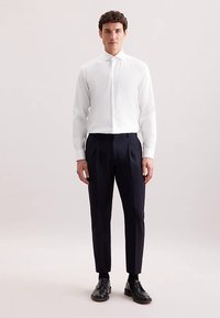 Camicia bianca a coste con colletto appuntito, abbinata a pantaloni blu scuro aderenti. Il modello mostra eleganti scarpe nere da uomo.