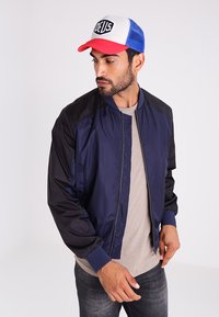 Chaqueta de bombardero azul marino con mangas negras, puños acanalados y cierre de cremallera. Llevada con una camiseta beige y una gorra de béisbol roja y azul.
