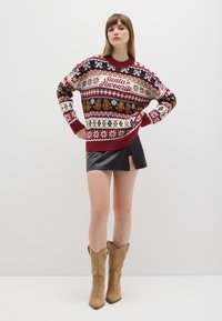 Red and white knitted sweater with festive patterns, including teddy bears and snowflakes, worn with a black leather mini skirt and beige boots.