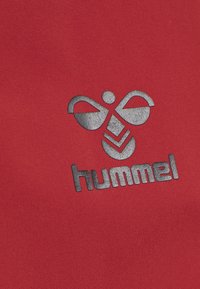 Rode stof met een zilver logo waarop een gestileerd figuur en chevronpatronen zijn afgebeeld, met de tekst "hummel" eronder.