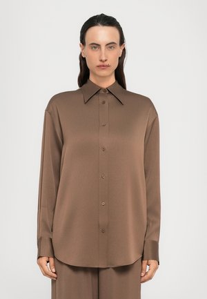 Woman wearing a loose brown long-sleeve button-up shirt with wide collar, standing straight against a plain light background.