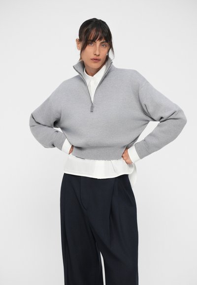 Pulover scurt gri cu guler înalt, fermoar complet și mâneci largi, suprapus peste o cămașă albă și asortat cu pantaloni evazați bleumarin.