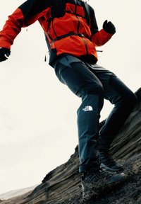 Crne planinarske hlače s sivim akcentima i logotipom The North Face, uparene s crnim robusnim obućom, postavljene na stjenovitom terenu.