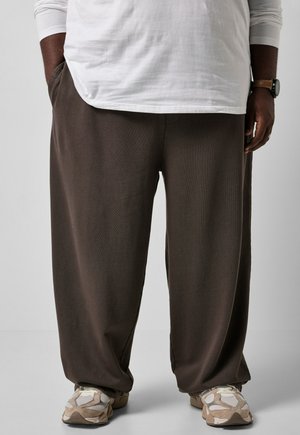 Person iført løstsiddende mørkebrune bukser, hvid skjorte med lange ærmer, beige og hvide sneakers, med den ene hånd i lommen og et ur på håndleddet.