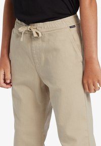 Beige katoenen broek met een elastische tailleband, trekkoord en zijzakken. De stof lijkt zacht met een subtiele textuur.