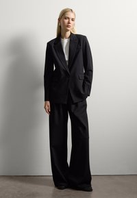 Blazer negro ajustado con solapas de muesca, cierre de un solo botón y bolsillos de parche, combinado con pantalones anchos negros y zapatos planos.