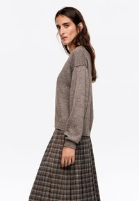 Brązowy dziergany sweter o pudełkowym kroju, z prążkowanymi mankietami, zestawiony z szarą plisowaną spódnicą w kratę, z pomarańczowymi i niebieskimi akcentami.