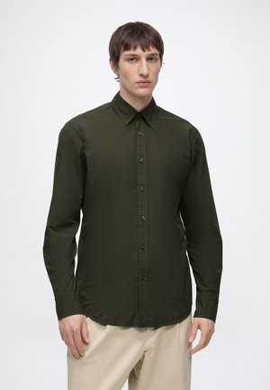 Homme portant une chemise olive à manches longues boutonnée et un pantalon beige clair, debout devant un fond blanc uni.