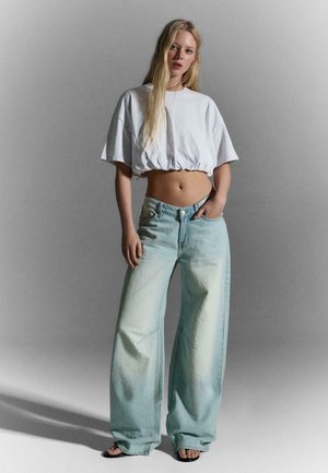 Femeie cu păr lung și blond poartă un tricou alb scurt, blugi largi de culoare albastru deschis și sandale negre, stând pe un fundal gri simplu.