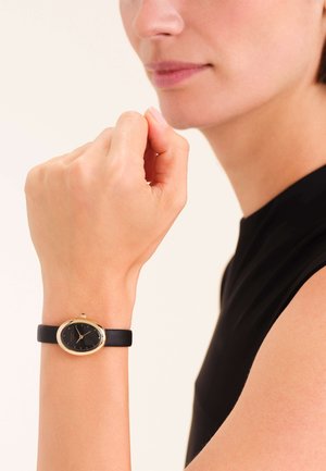 Montre à bracelet en cuir noir avec un boîtier ovale doré et un cadran noir, présentant des marqueurs d'heure minimalistes et une petite couronne.
