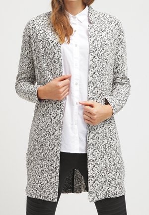 Person iført en lang, sort-hvid mønstret cardigan med åben front over en hvid skjorte med knapper og sorte bukser.