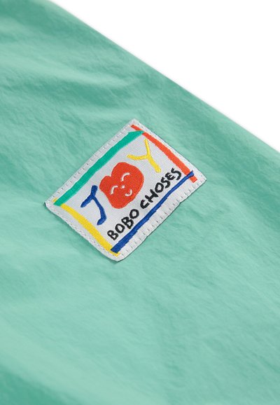 Hellgrüner Stoff mit einer strukturierten Oberfläche, versehen mit einem bunten gestickten Etikett, das ein lächelndes Gesicht und den Text "BOBO CHOSES" zeigt.