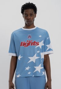 Άνδρας με μπλε αθλητική φανέλα με λευκά αστέρια και κόκκινο κείμενο "low lights" που στέκεται μπροστά σε απλό φόντο.