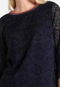 Top in pizzo navy con motivo floreale, caratterizzato da maniche a tre quarti e scollatura rotonda con rifiniture rosse e bianche. Texture morbida.