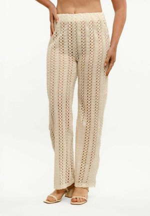 Beige gehaakte gebreide broek met hoge taille en rechte pijpen, gedragen met beige open-toe blokhakken sandalen, hand van het model rustend op de heup.