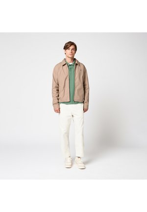 Jeune homme debout portant une veste beige, un polo vert, un pantalon blanc et des baskets blanches sur un fond clair uni.