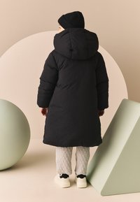Cămașă căptușită neagră cu o glugă mare, asortată cu pantaloni gri matlați și adidași albi, pe un fundal cu forme geometrice minimaliste.