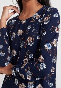 Blusa floreale blu navy con maniche lunghe, leggeri motivi floreali in bianco, arancione e blu, colletto rotondo e tessuto texturizzato.