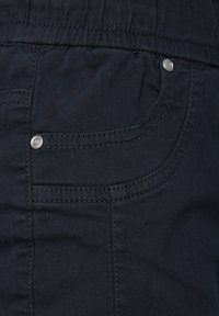 La tela de denim azul oscuro presenta un bolsillo lateral con costuras en contraste y detalles de remaches metálicos, mostrando una textura suave y un diseño estructurado.