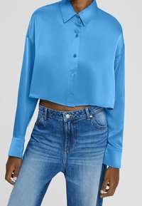 Modello che indossa una camicia corta blu brillante con bottoni e maniche lunghe, abbinata a jeans blu sbiaditi a vita alta.