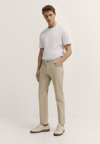 Weißes T-Shirt mit kurzen Ärmeln, beigefarbene gerade Hosen und hellbeige Sneaker mit braunen Sohlen. Einfaches Design, glatte Textur, neutrale Farben.