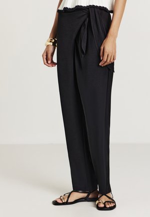 Schwarze weite Hosen mit einem Bund aus glattem Stoff, mit einer Schleife in der Taille. Kombiniert mit flachen schwarzen Sandalen mit goldenen Akzenten.