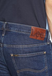 Tmavě modré džíny z denimu s hnědou koženou štítkem s nápisem "Lee" na zadním pásku. Obsahují více kapes a žluté švy jako akcenty.