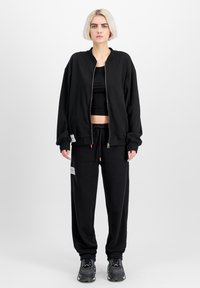 Crna bomber jakna sa zatvaračem preko crnog crop topa, u kombinaciji s crnim trenirkama. Sadrži elastične manžete i vidljive oznake brenda.