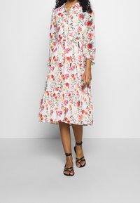 Blumiges Midikleid mit langen Ärmeln, durchgehender Knopfleiste, weißem Hintergrund, pinken und roten Blumen, sch tiered Rock, kombiniert mit schwarzen Riemchensandalen.