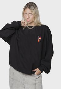 Sudadera negra de gran tamaño con un acabado texturizado, que presenta un pequeño gráfico bordado de un encendedor con llamas en el lado izquierdo del pecho.