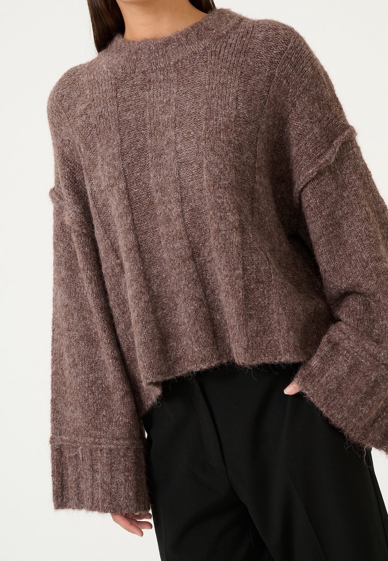 Femme portant un pull marron ample et texturé avec de larges manches, associé à un pantalon noir sur un fond uni.