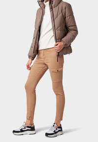 Chaqueta de plumas acolchada marrón con capucha, suéter de cuello alto blanco, pantalones cargo beige con bolsillo lateral y zapatillas negras/blancas.