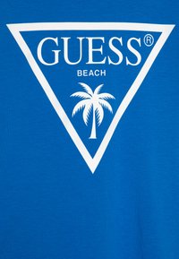 Plava pamučna majica s bijelim trokutastim grafičkim prikazom koji u sredini sadrži tekst "GUESS BEACH" i siluetu palme.