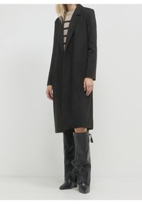 Femme portant un long manteau noir par-dessus un pull beige à rayures et des bottes hautes en cuir noir, debout devant un fond uni.