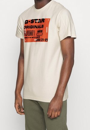 Beige bomulds t-shirt med et iøjnefaldende orange og sort grafisk tryk på fronten, med en afslappet pasform og korte ærmer.