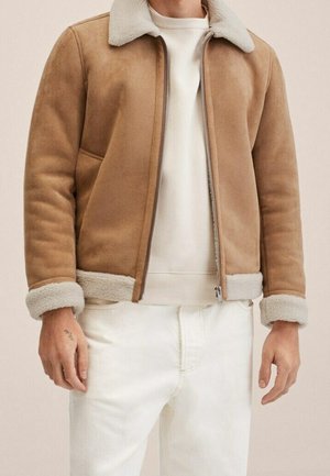 Man in tan lammyjas met crèmekleurige fleece kraag en manchetten, gedragen over een wit overhemd en witte broek, staand met ontspannen armen.