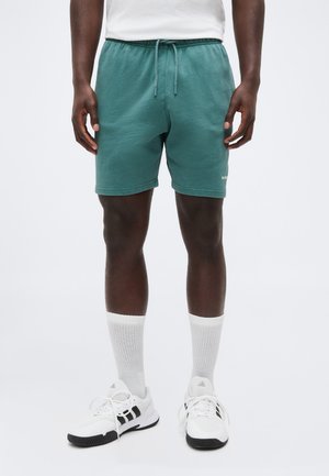 Mann iført turkise shorts med snøring, hvite sokker til midt på leggen og hvite joggesko med svarte striper, stående mot en ensfarget bakgrunn.