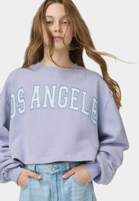 Beskuren lavendelfärgad sweatshirt med "LOS ANGELES" tryckt i ljusblått, med långa ärmar och rund halsringning. Kombinerad med högmidjade jeans.