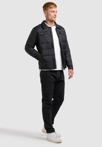 Jeune homme debout portant une doudoune noire, un t-shirt blanc, un jean noir et des baskets blanches devant un fond gris uni.