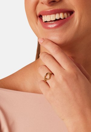 Smiling woman touching chin with hand wearing gold heart-shaped ring, light pink top visible on shoulder.