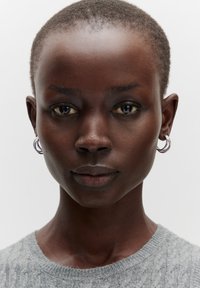 Model mit kurzem, dunklem Haar und Creolenohrringen, trägt einen hellgrauen Strickpullover. Hintergrund ist schlicht weiß. Hautfarbe ist dunkel.
